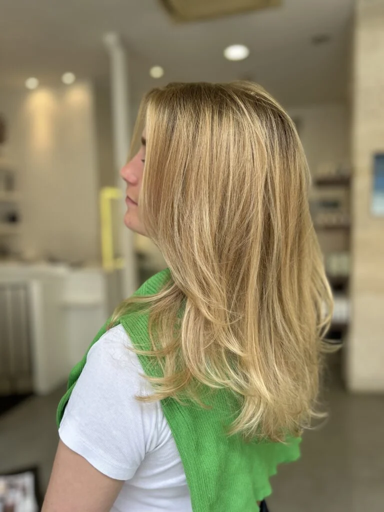 Coiffure femme au cheveux blond dans un salon de coiffure . Elle est de profile est l'on voit bien sa frange rideau. Elle est habille en blanc avec un pull vert anis sur ses épaule